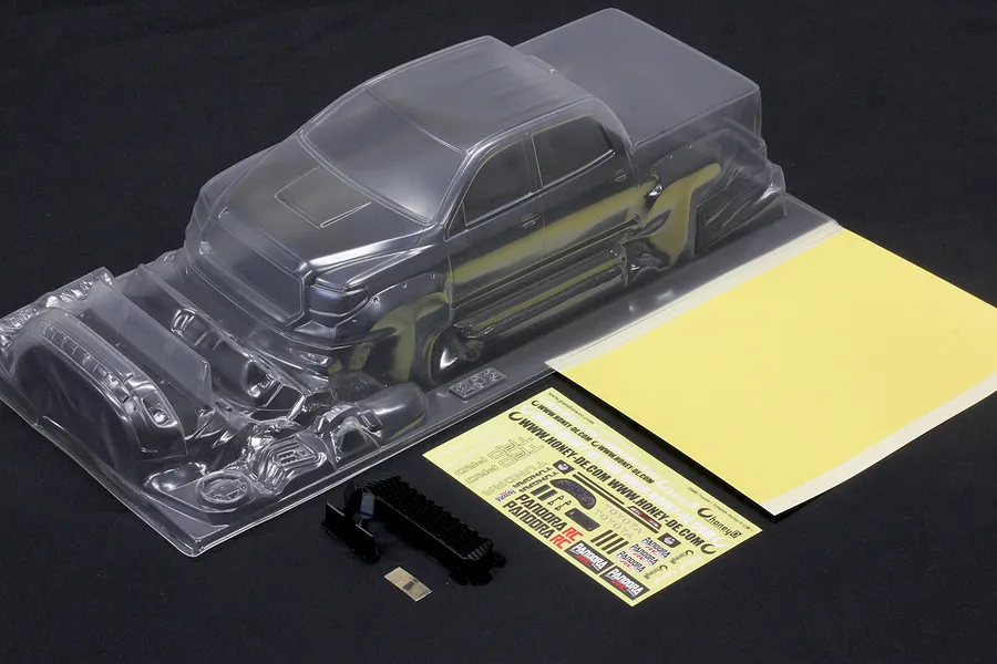 Unpainted clear polycarbonate RC truck body shell with separate parts, decal sheet, and window mask on a dark background.