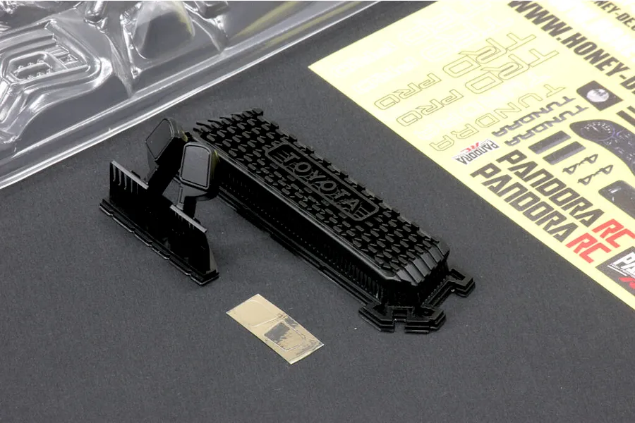 Unpainted black plastic RC truck accessories including a grille, mirrors, and a decal sheet on a dark surface.