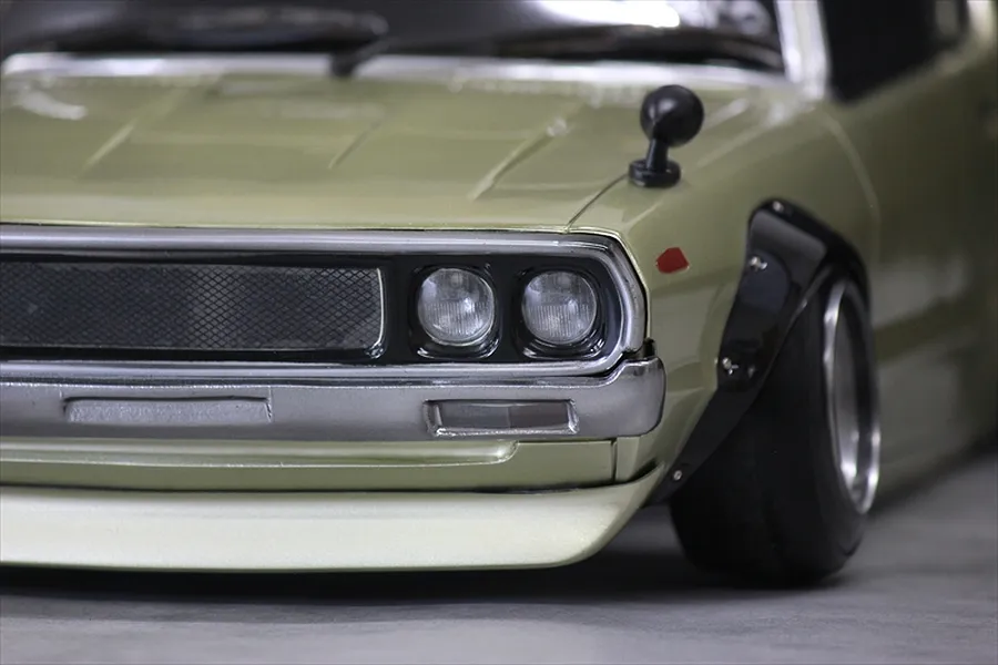 Close-up studio view of the front corner of a gold RC body shell showing headlights, grille, fender flare, and bumper.