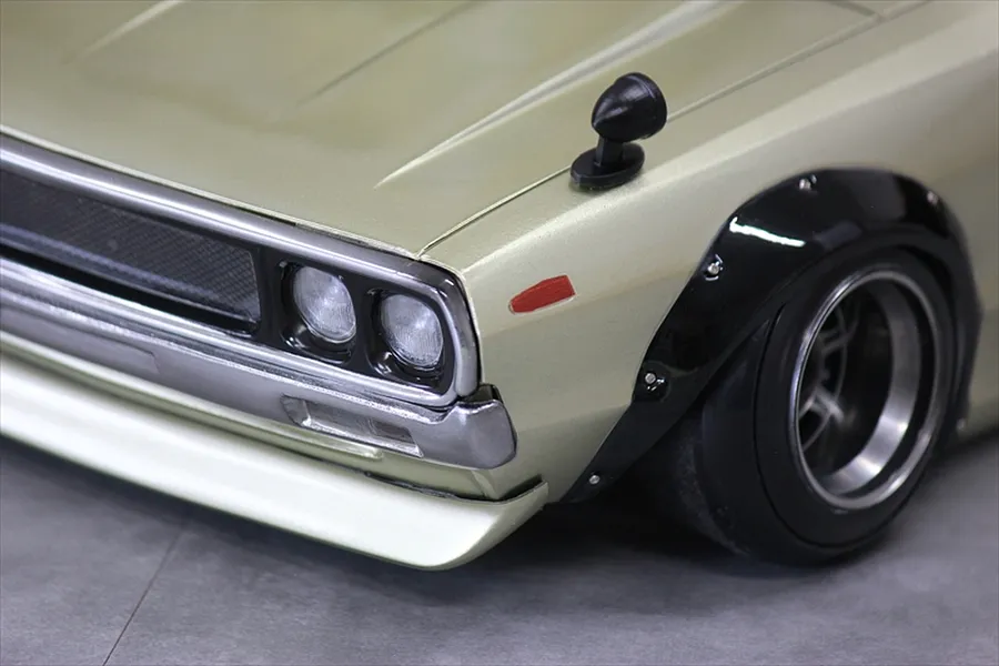 Close-up of the front left corner of a gold RC body shell showing headlights, grille, fender flare, and wing mirror.