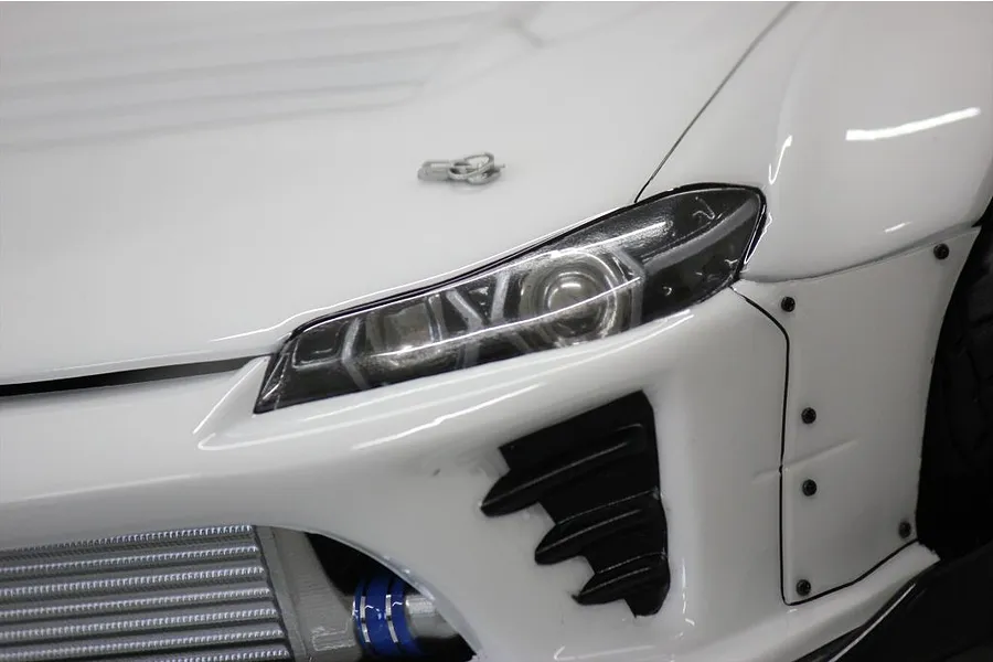 Close-up of the front corner of a white RC drift car body showing the headlight, front bumper, intercooler, and body pins.