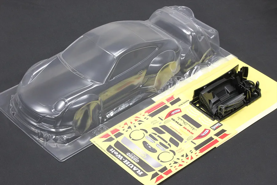 Unpainted clear polycarbonate RC body shell of a Porsche 997 with wide fenders, accompanied by decal sheets and black plastic parts on a dark surface.