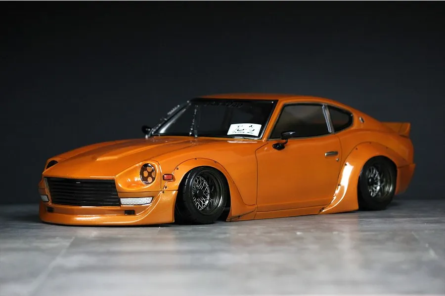 An orange custom S30 Fairlady Z RC drift car body shell with wide fender flares and black mesh wheels parked against a dark background.