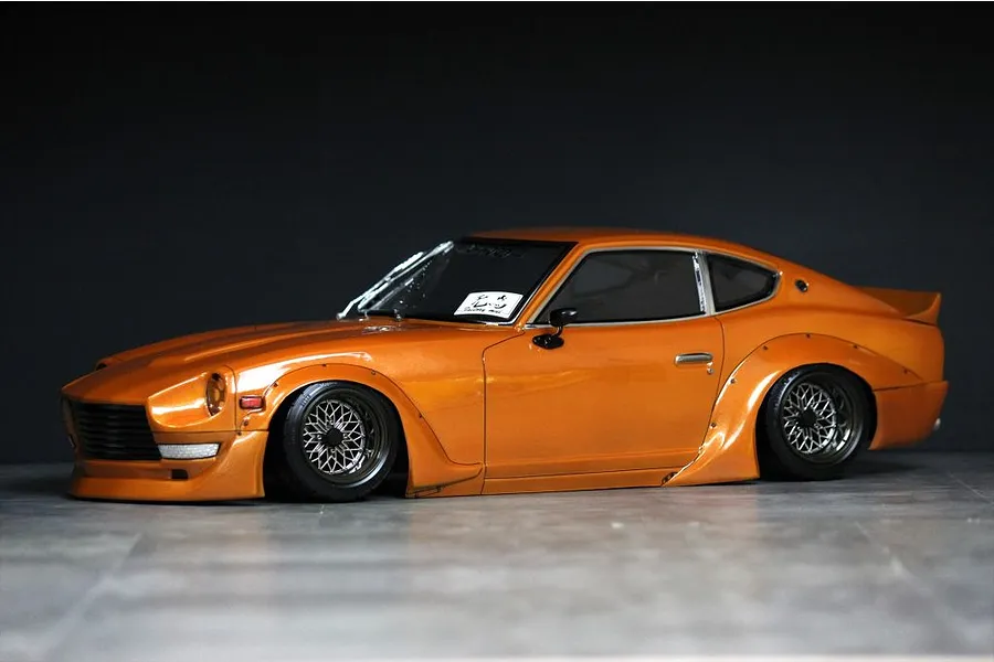 Custom painted orange S30 Fairlady Z RC drift car body shell with wide fender flares and wire mesh wheels on a dark studio background.