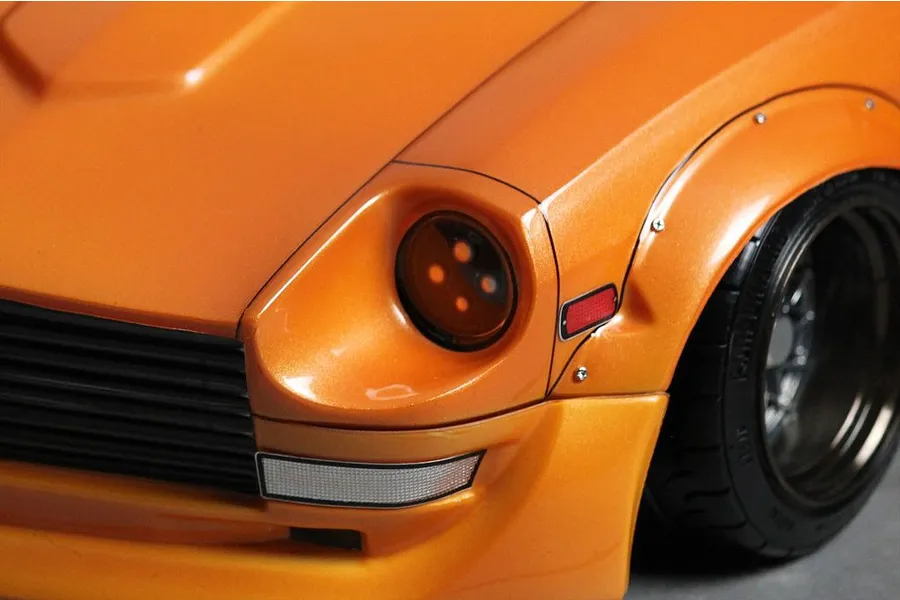 Close-up of the front corner of an orange RC car body shell, showing the headlight, grille, fender flare, and wheel.