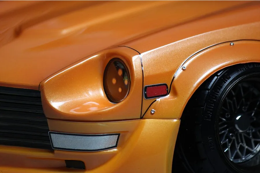 Close-up of metallic orange RC model car body featuring front headlight bucket, fender flare with visible screws, and black multi-spoke wheel.