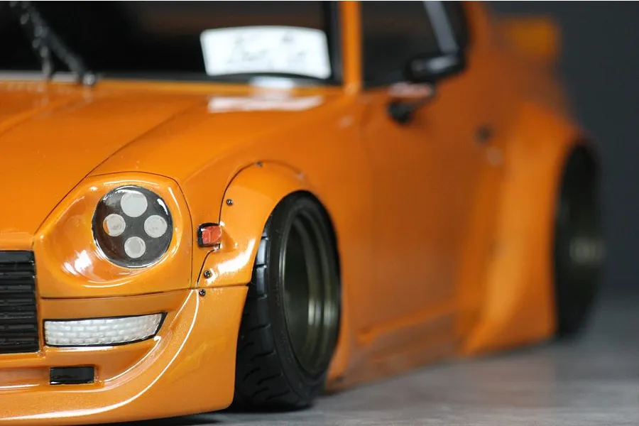 Close-up of an orange RC car body shell with flared fenders, custom wheels, and detailed headlights on a dark background.