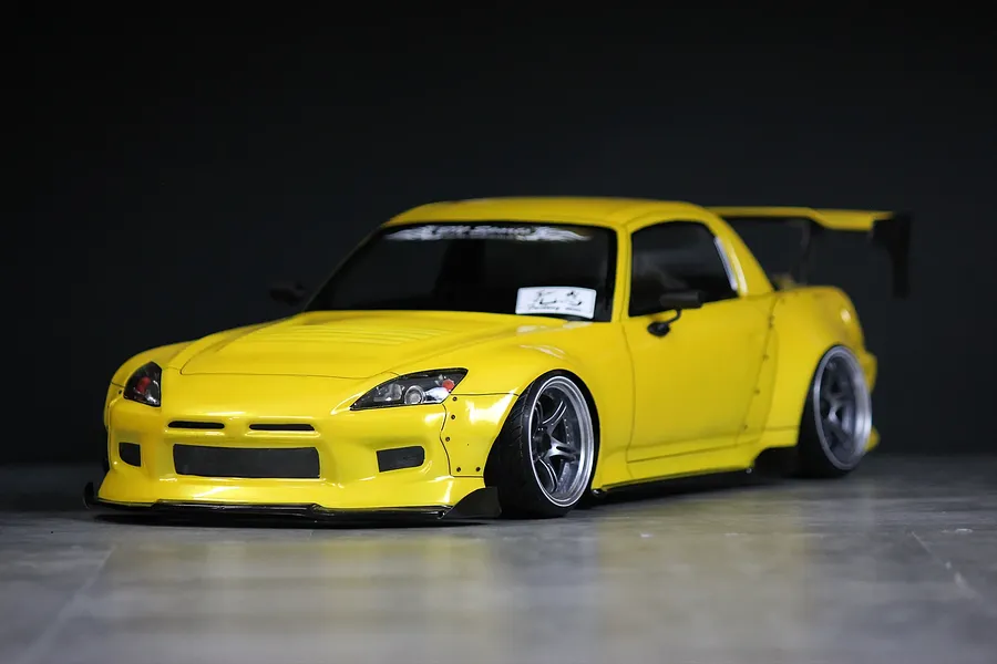 Custom painted yellow Honda S2000 RC drift car body shell with aftermarket aero kit, wide fenders, and black wheels against a dark background.