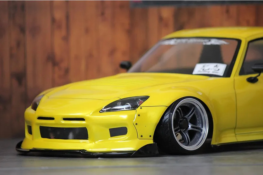 Close-up lifestyle shot of a yellow RC drift car body shell mounted on a chassis with custom wheels against a wooden background.