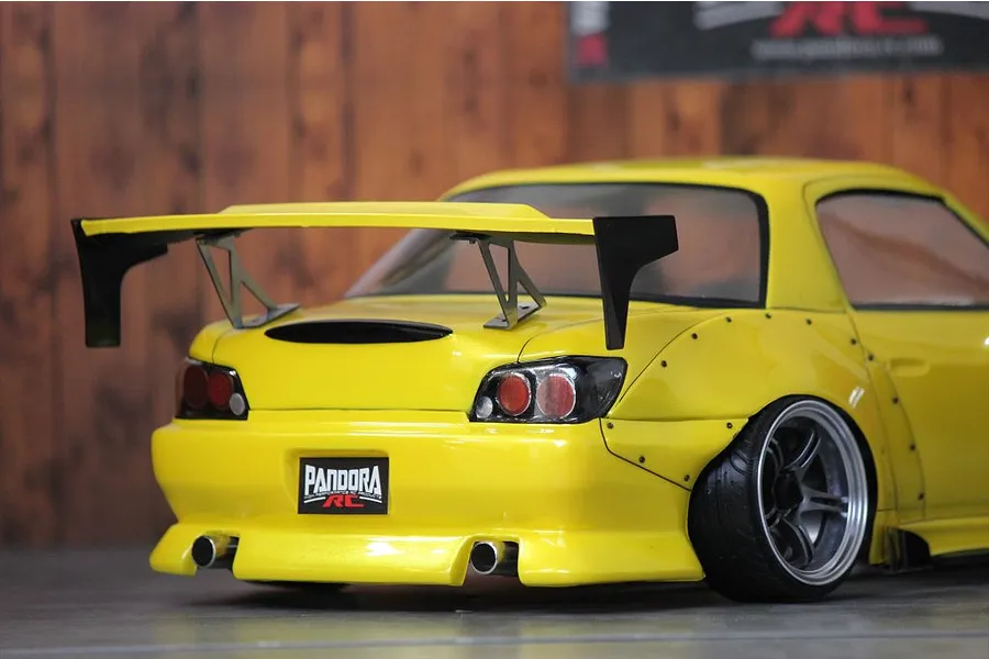 Rear view of a yellow RC drift car body with a large black wing, rear diffuser, and Pandora RC license plate against a wooden wall background.