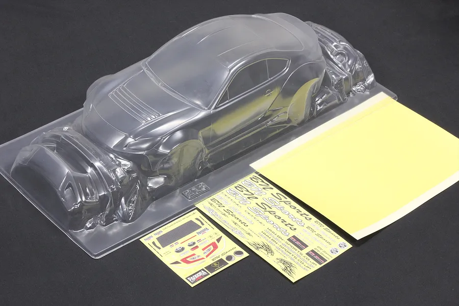 Unpainted clear polycarbonate RC car body shell with accessory parts, decal sheets, and window masks on a dark surface.