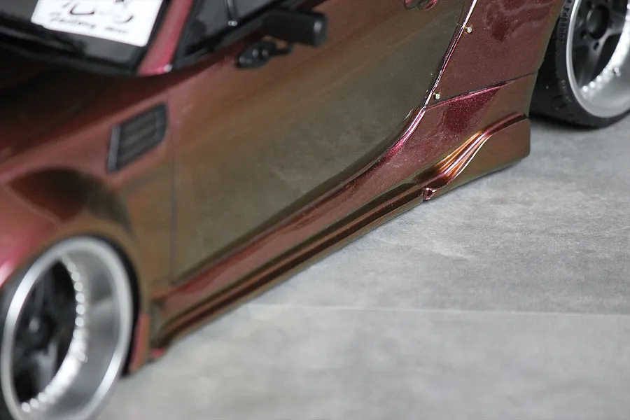 Close-up of the side profile and skirt of a metallic maroon RC drift car body with widebody fender flares and deep-dish wheels.