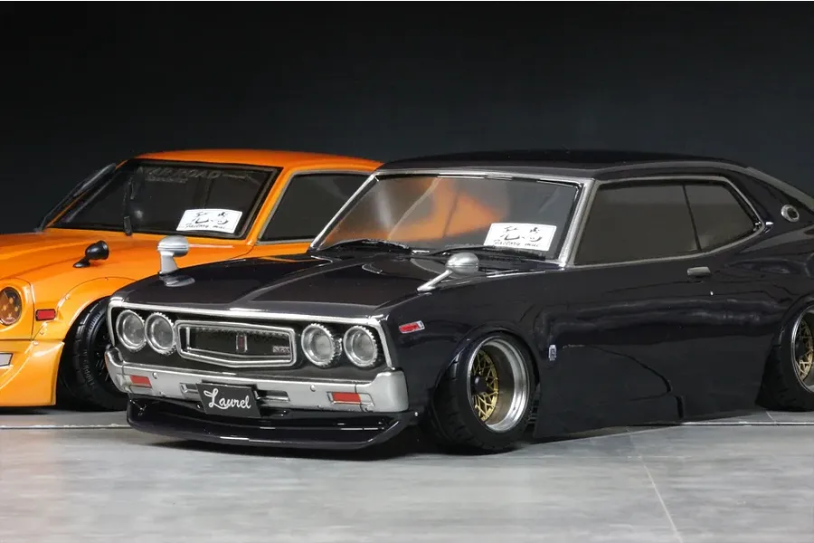 A painted C130 Laurel RC body shell in dark metallic grey displayed next to an orange one on a studio floor.