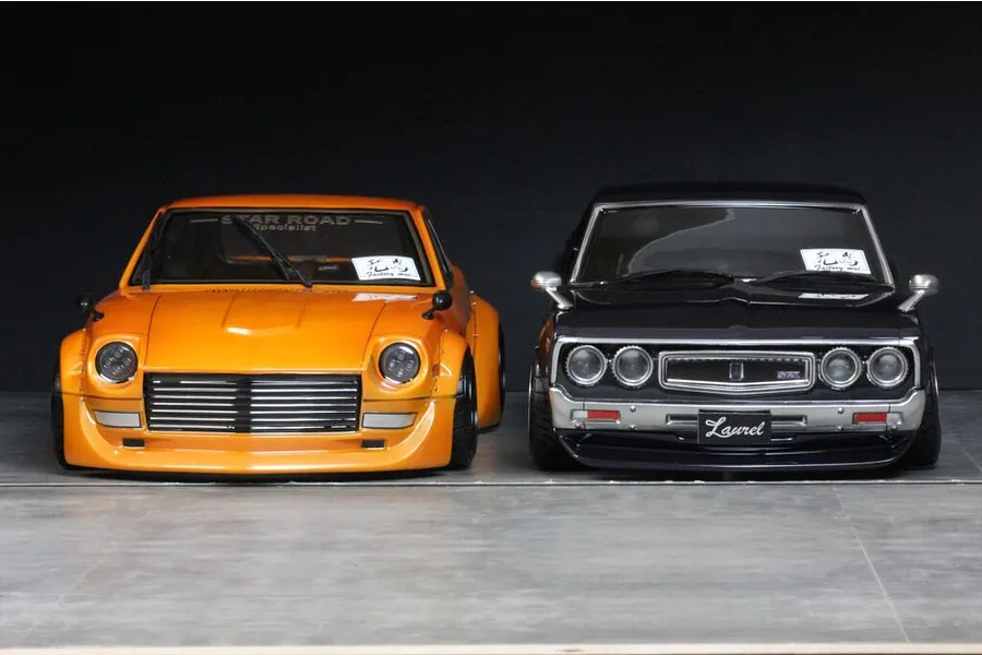 Two custom RC drift car bodies displayed side-by-side on a grey surface against a dark background, showing an orange model and a dark grey model.