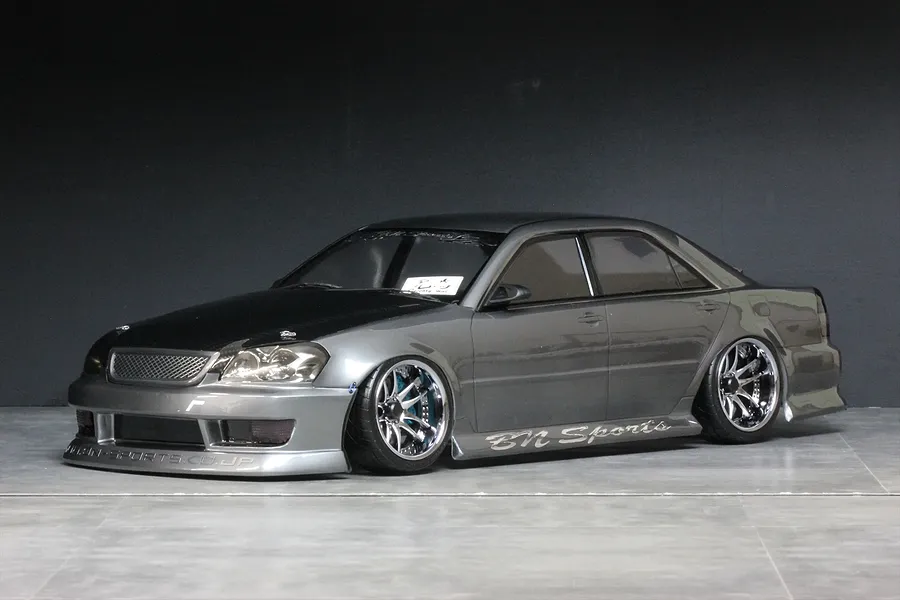 Studio photo of a grey Toyota Mark 2 JZX110 BN-Sports RC drift car body shell mounted on a chassis with chrome deep dish wheels.