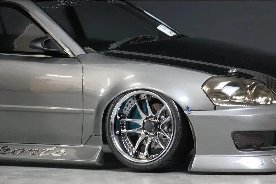 Close-up of a silver RC drift car body showing the front wheel, chrome multi-spoke rim, and black hood.