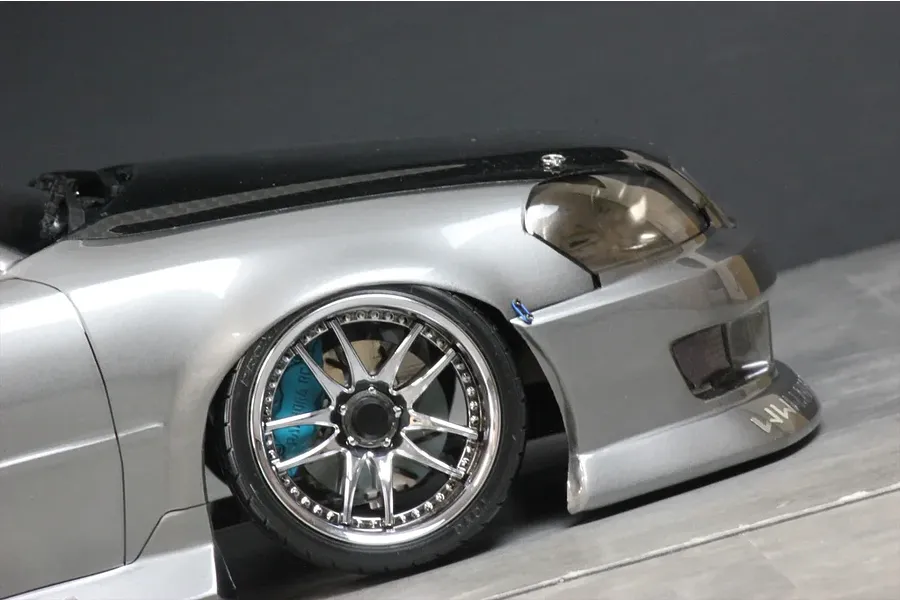 Close up on the front wheel and fender of a silver RC drift car body with chrome rims and blue brake calipers.