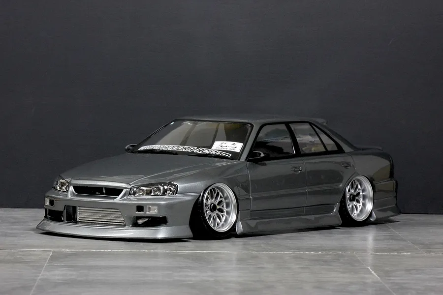 Studio product shot of a grey ER34 Skyline 4-door RC drift car body shell mounted on a chassis with custom wheels against a dark grey background.