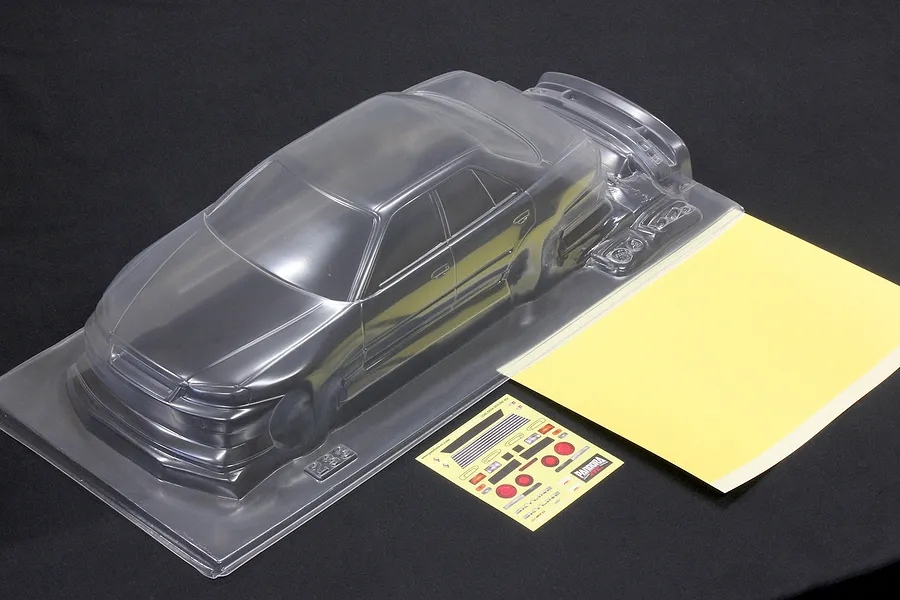 Unpainted clear polycarbonate RC car body shell with a mask sheet and sticker sheet for the ER34 Skyline 4Dr on a dark surface.