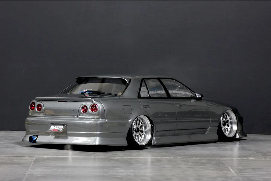 Rear three-quarter view of a grey Pandora RC ER34 Skyline 4-door drift RC car body shell with custom wheels and exhaust tip on a studio floor.