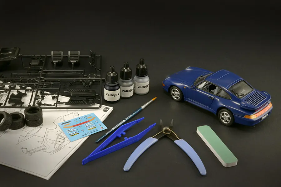 An assembled blue Porsche 911 model car alongside plastic parts sprues, tools, paint bottles, and instruction sheet on a dark background.