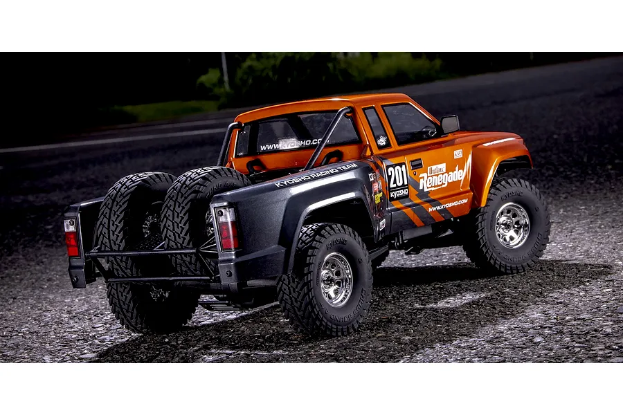 Kyosho Outlaw Renegade 2RSA RC short course truck parked outdoors on asphalt, showing its rear-mounted spare tires and orange and grey body.