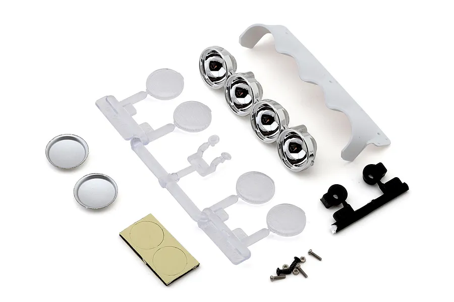 RC car scale accessory parts including chrome light buckets, clear plastic tree, white wing, screws, and adhesive pads on a white background.