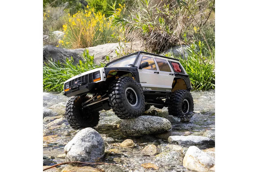 Axial SCX10 III Jeep Cherokee XJ white RC rock crawler driving over wet river rocks in an outdoor natural setting.