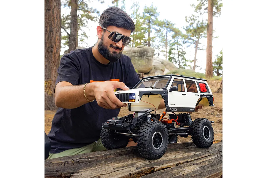 Man adjusting the white body of an Axial SCX10 III Jeep Cherokee XJ RC rock crawler outdoors on a log.