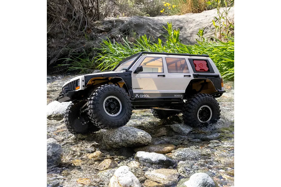 Axial SCX10 III Jeep Cherokee XJ RC rock crawler driving over rocks and through a shallow stream in an outdoor setting.