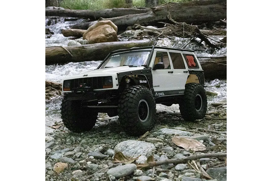 Axial SCX10 III Jeep Cherokee XJ RC rock crawler driving on rocky ground beside a stream outdoors.