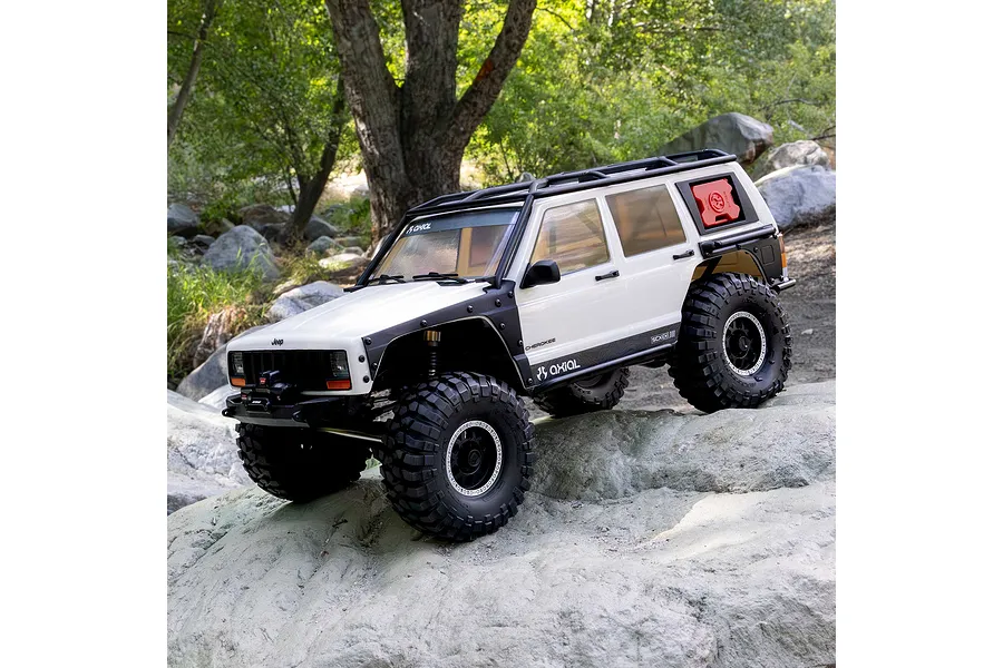 Axial SCX10 III Jeep Cherokee XJ white RC rock crawler driving outdoors on rocky terrain among trees.