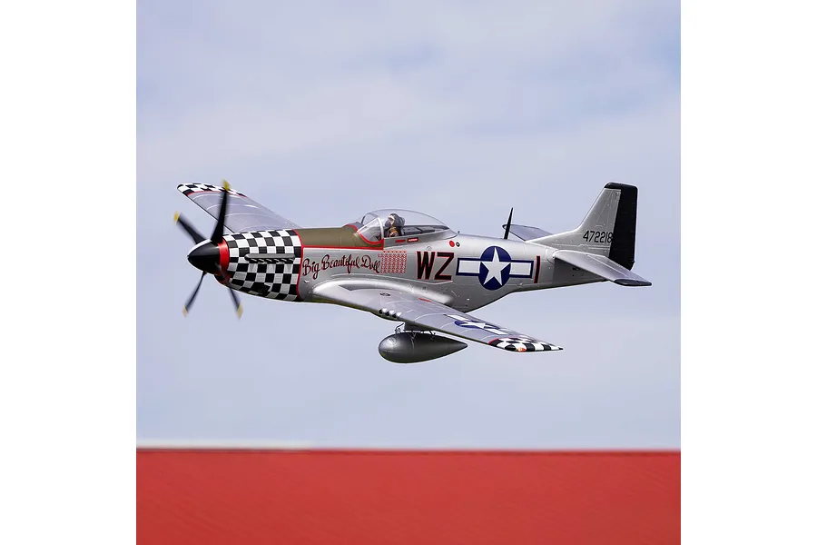 E-Flite P-51D Mustang Big Beautiful Doll RC airplane in flight against a blue sky with a red roof below.