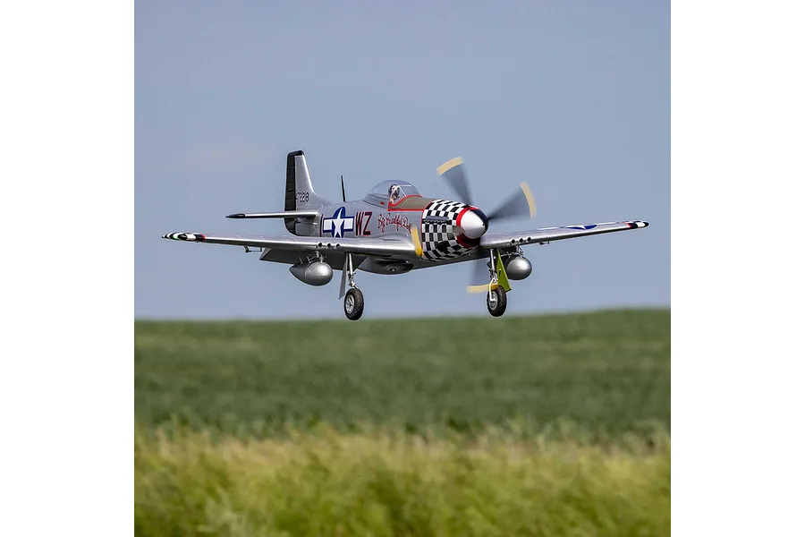 E-Flite P-51D Mustang Big Beautiful Doll RC fighter plane in flight over grassy fields with landing gear deployed.