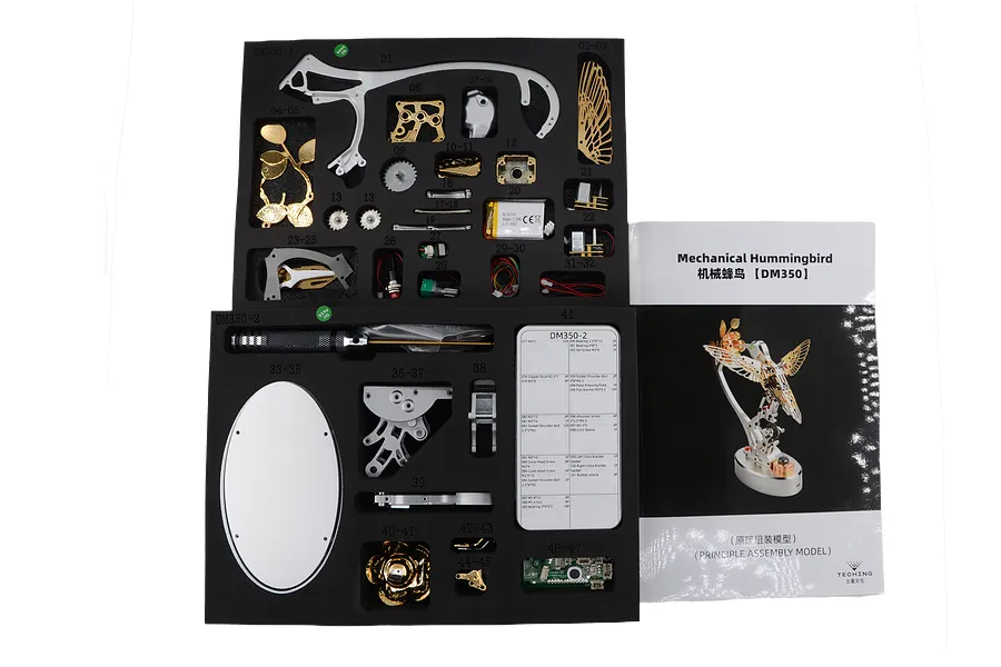 Overhead studio view of the Teching DM350 Mechanical Hummingbird parts laid out in black foam trays next to its instruction manual.