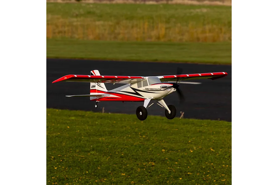 E-Flite Turbo Timber Evolution 1.5m RC plane in flight over an airfield with a red, white, and black trim scheme.