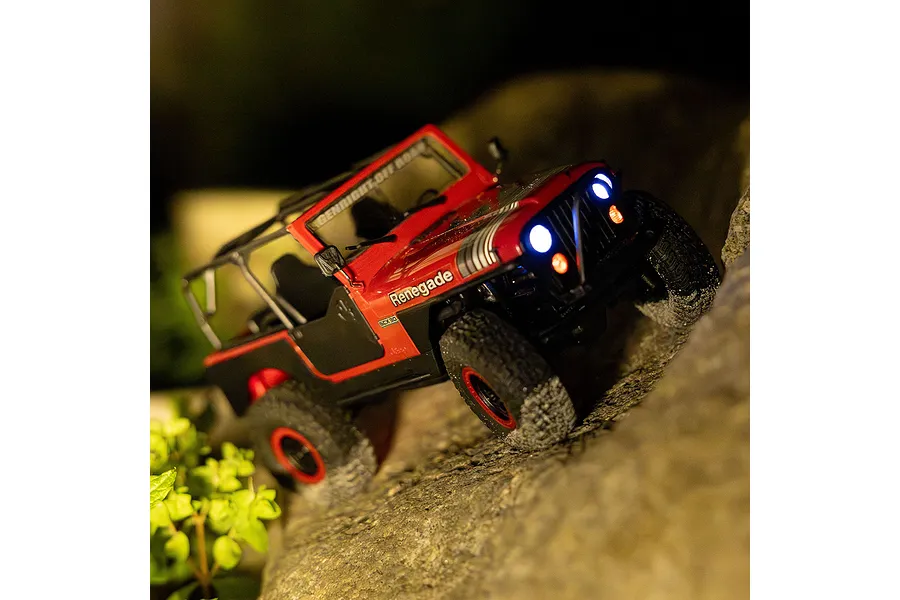 Red Axial SCX30 Jeep CJ-7 Renegade RC rock crawler driving on rocks at night with working headlights lit up.