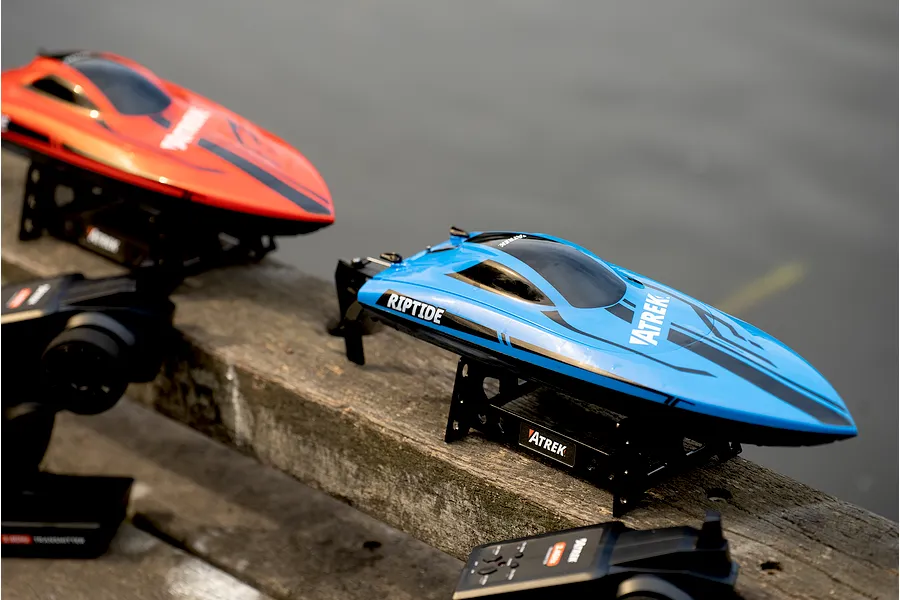 Blue Atrek Riptide RC speed boat and transmitter on a wooden surface with a red boat in the background.