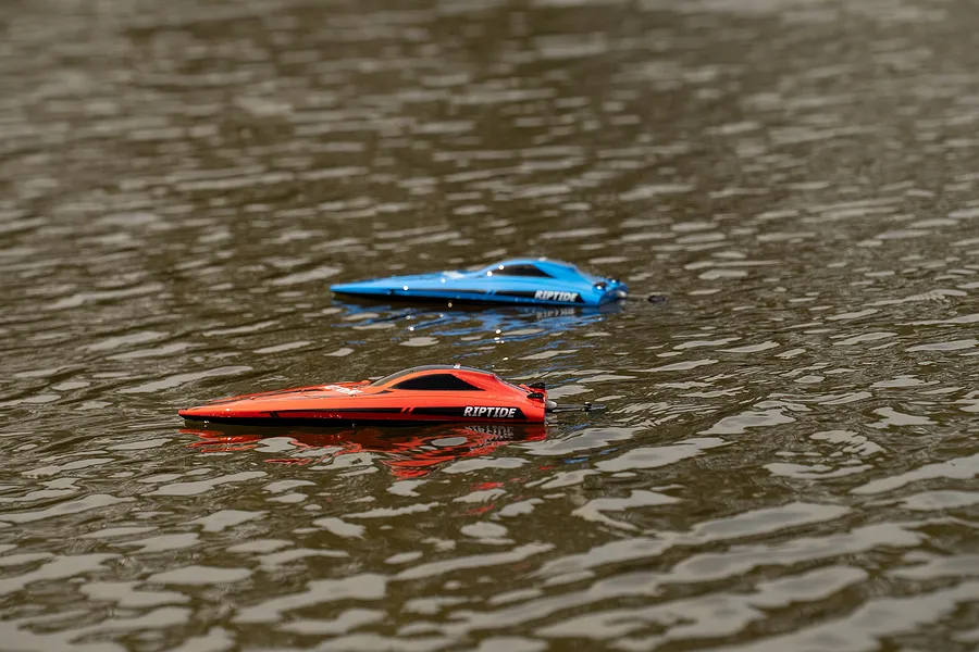 Two Riptide RC speed boats, one red and one blue, floating on rippling outdoor water.