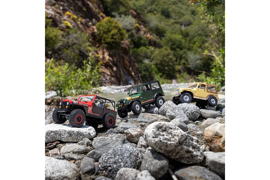 Three Jeep RC rock crawlers in red, green, and yellow navigating rocky terrain near a stream.