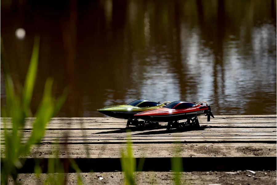 Two Atrek Rogue RC speed boats, one orange and one green, sitting on display stands on a wooden dock by the water.