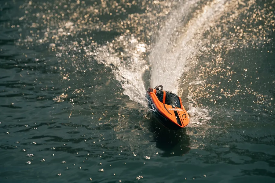 An orange and black RC speed boat cuts through open water, creating a dynamic spray and wake.