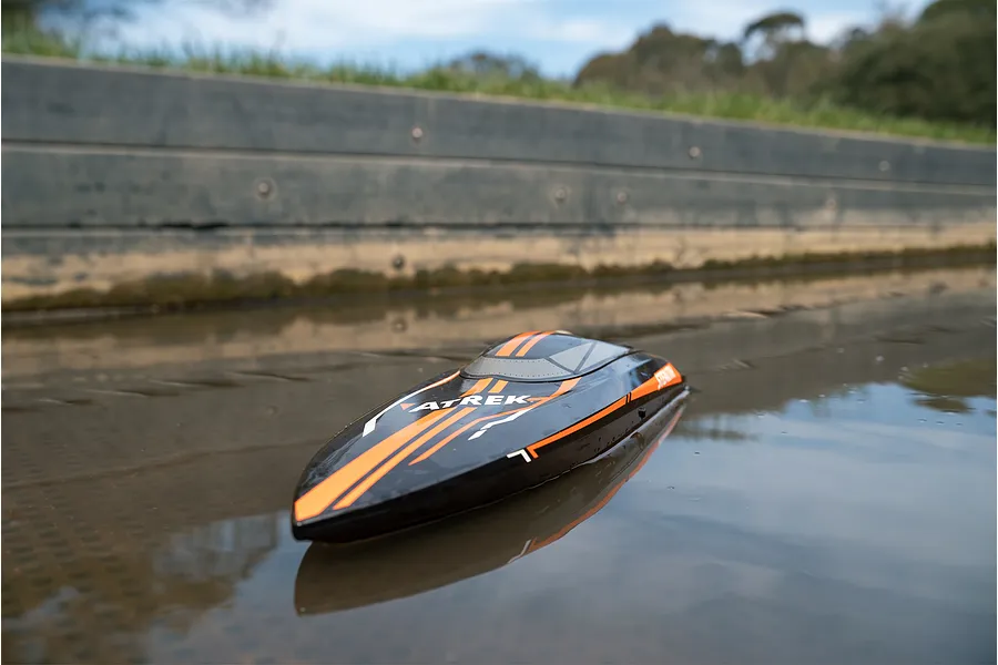Atrek Stealth black and orange RC speed boat floating in calm water near a concrete retaining wall outdoors.