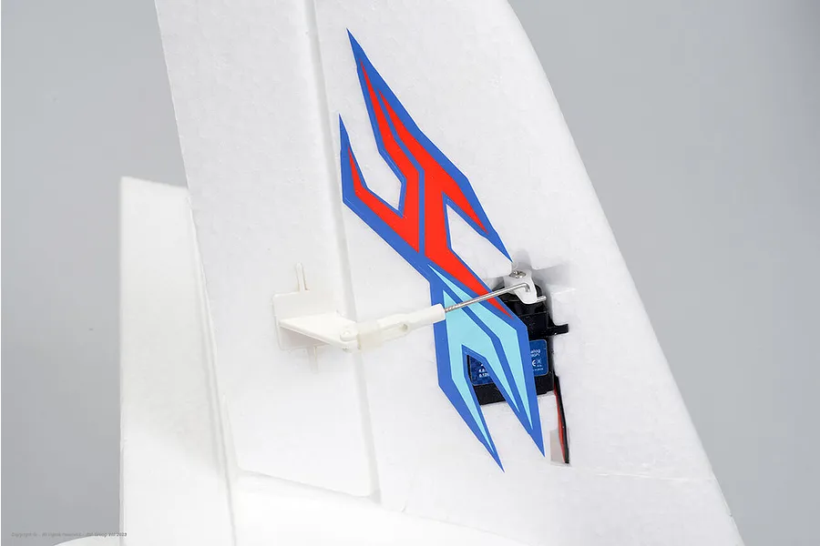Close-up of an RC plane tail section showing white foam vertical stabilizer, servo, control linkage, and red, dark blue, and light blue decals.