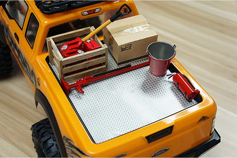 A silver diamond plate sheet installed in the bed of an orange RC truck, topped with scale accessories like a crate, box, bucket, and fire extinguisher.
