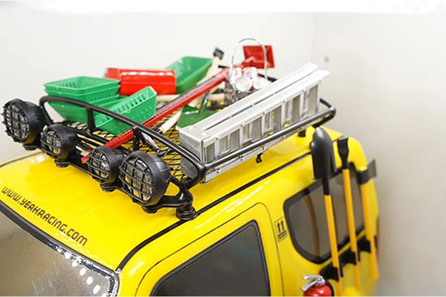 A silver aluminium scale ladder mounted on the roof rack of a yellow RC crawler vehicle, alongside other scale accessories like baskets and tools.