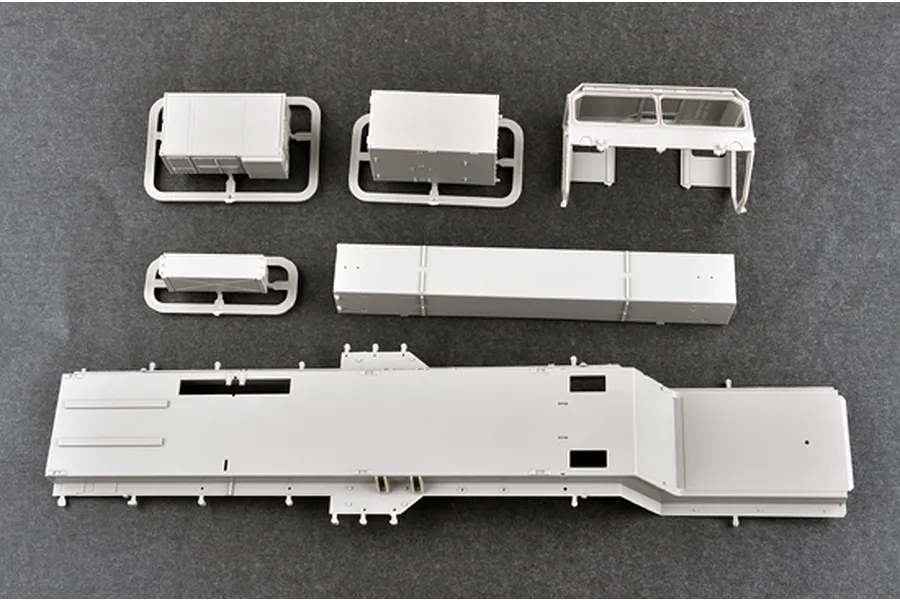 Unassembled grey plastic model kit parts for a military vehicle, including a large chassis, truck cab, and storage boxes on a dark surface.