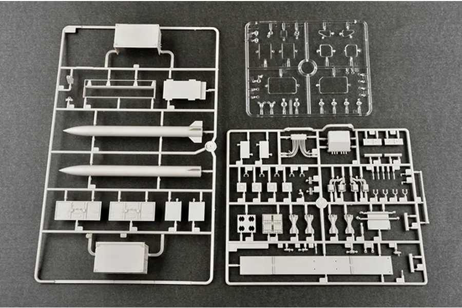 Three plastic model kit sprues containing grey and clear parts for a military vehicle and missile system on a dark grey surface.