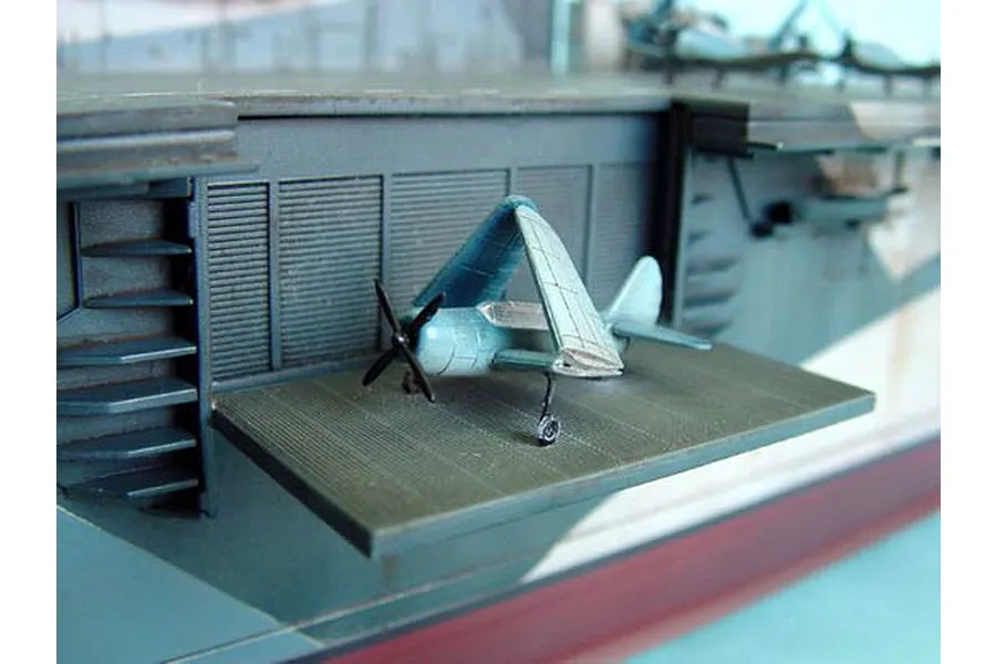 A close-up of a 1/350 scale model aircraft carrier deck with a small blue fighter plane featuring folded wings parked on an elevator platform.