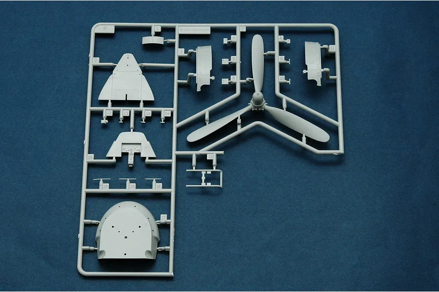 A grey plastic model kit sprue featuring a three-blade propeller and various aircraft fuselage parts on a blue background.